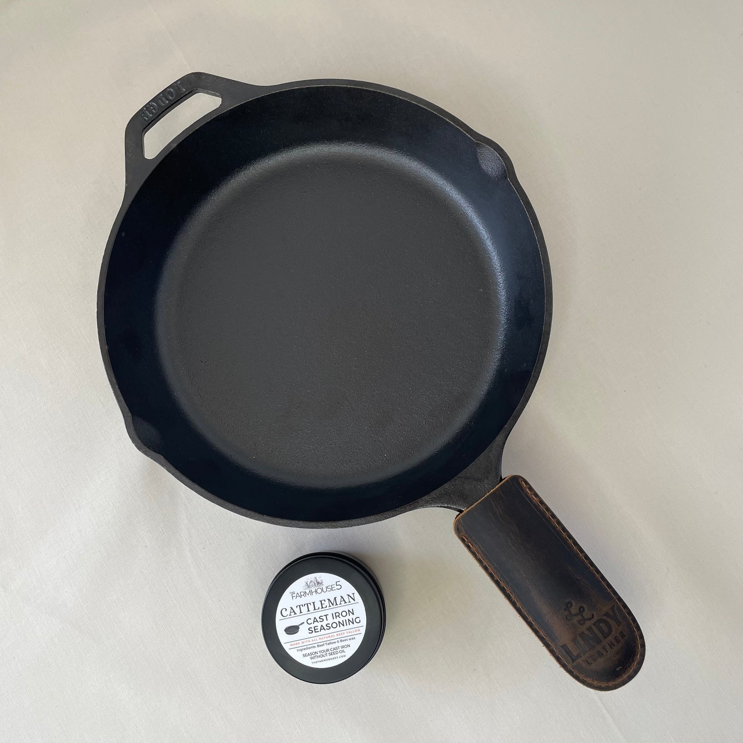 Made in USA cast iron skillet kit: Lodge 10.25” pre-seasoned skillet, Cattleman tallow seasoning tin, and Lindy leather handle cover on beige background