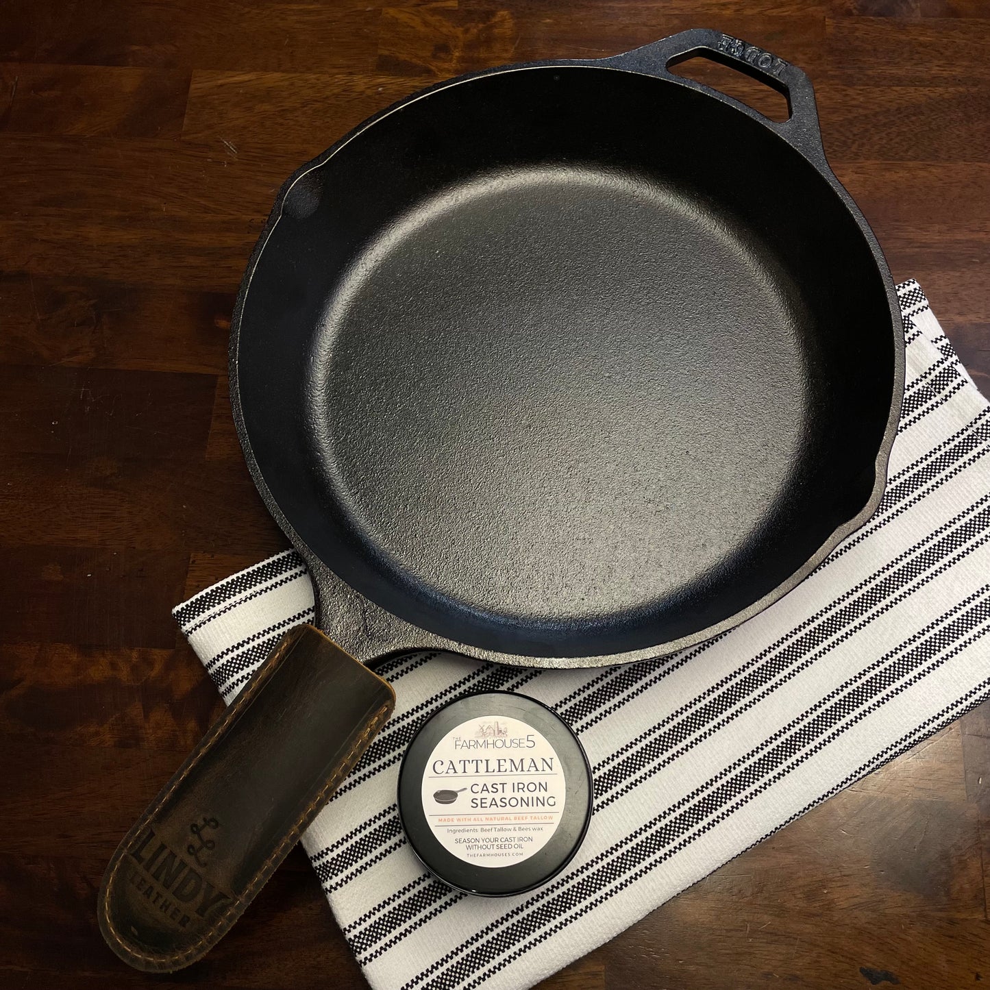 Made in USA cast iron skillet kit: Lodge 10.25” pre-seasoned skillet, Cattleman tallow seasoning tin, and Lindy leather handle cover on rustic wood surface