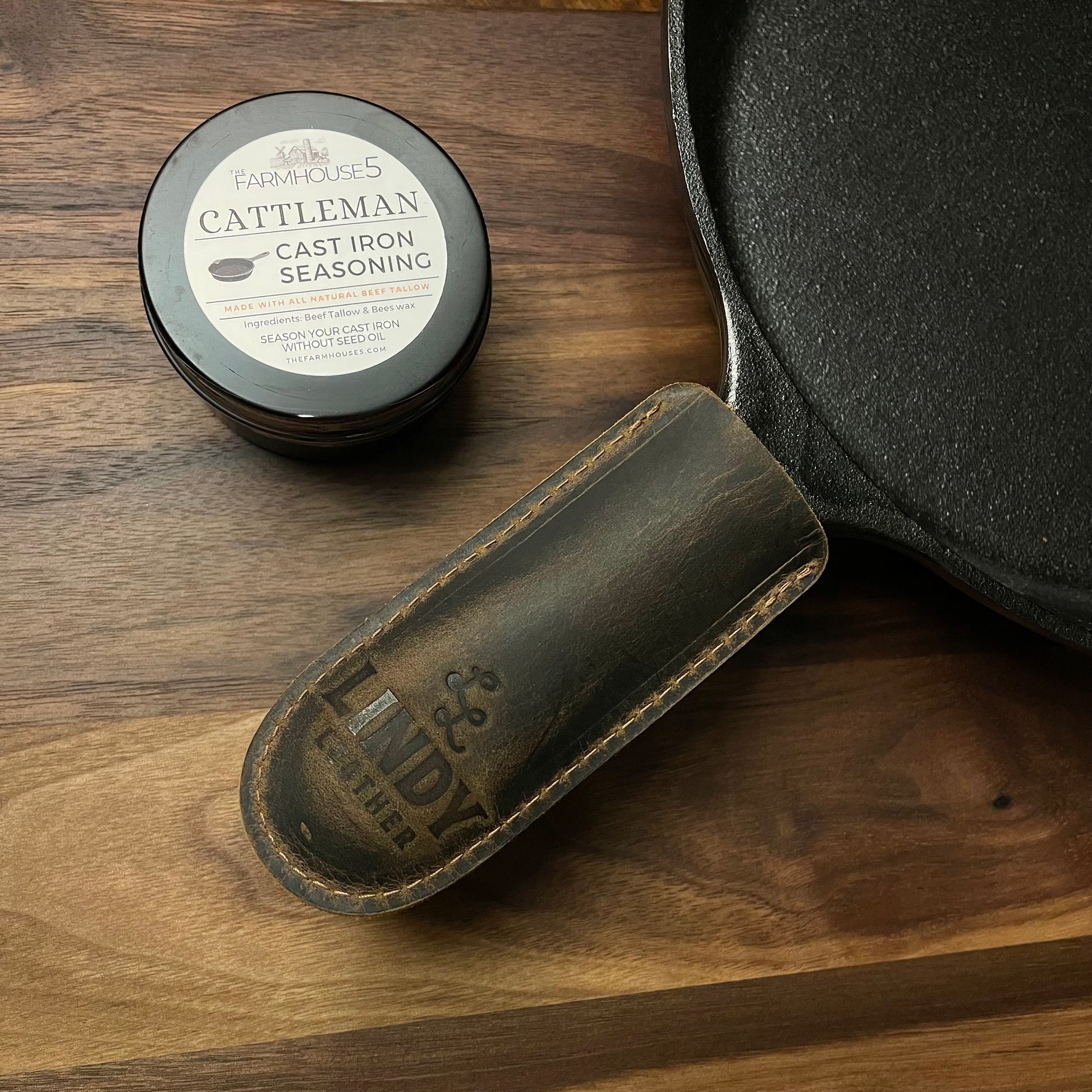 Made in USA cast iron skillet kit: Lodge 10.25” pre-seasoned skillet, Cattleman tallow seasoning tin, and Lindy leather handle cover on rustic wood surface